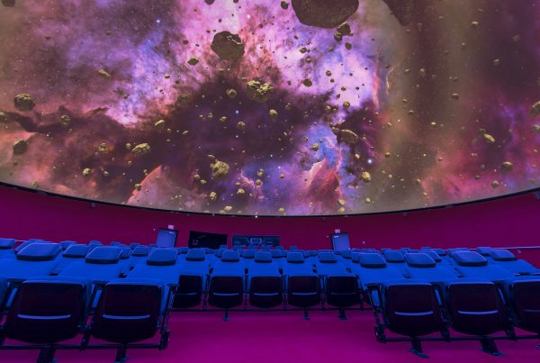 Interior of a planetarium theater. Seats are empty and a fulldome image of a nebula and asteroids is shown on the dome.